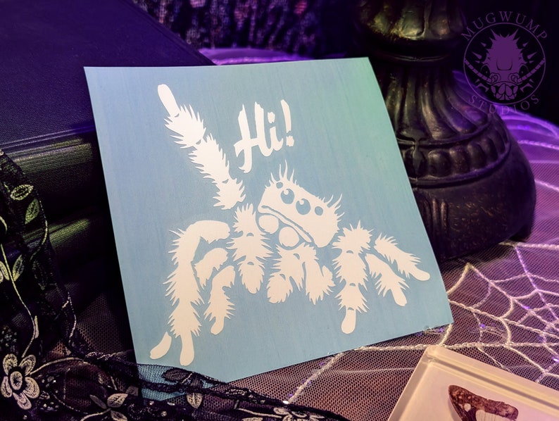 A photograph of a cute jumping spider decal. The jumping spider is looking up and raising one leg to wave a greeting. He says 'Hi!' The photograph is of the decal sitting top of a spiderweb lace tablecloth.