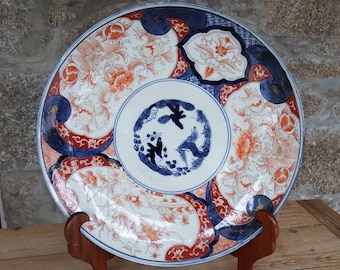 Może przedstawiać: Duży, okrągły, biały talerz porcelanowy z niebieskim i czerwonym wzorem kwiatowym. Talerz stoi na drewnianym stojaku.