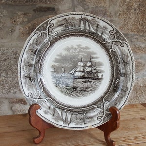 May include: A vintage ceramic plate with a black and white image of a sailing ship on the ocean. The plate has a rope design around the edge and is on a wooden stand.