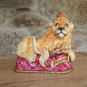 Könnte beinhalten: Eine Keramik-Löwenfigur mit einer goldenen Krone, die auf einem roten und goldenen Kissen sitzt. Der Löwe hat einen königlichen Ausdruck und hält ein goldenes Zepter in seiner Pfote.