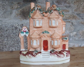 May include: A ceramic house figurine with a brown and white color scheme. The house has a red door and green ivy details. The figurine is sitting on a wooden surface.