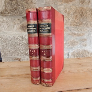 May include: Two vintage books with red leather covers. The spines display "ENGLISH ILLUSTRATED MAGAZINE" with "VOL. I" and "VOL. II" in gold. The books are on a wooden surface, set against a stone wall.