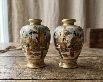 Może przedstawiać: Para małych ceramicznych wazonów o kremowej podstawie z misternie ręcznie malowanymi scenami przedstawiającymi postacie w tradycyjnych strojach. Wazony mają wąską szyjkę, rozszerzany wylot oraz złoconą podstawę z geometrycznym wzorem.