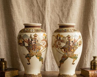 Może przedstawiać: Dwie kremowe wazony ceramiczne z misternymi, ręcznie malowanymi ilustracjami wojowników samurajskich w tradycyjnych zbrojach, ozdobione złotymi, czerwonymi i zielonymi detalami oraz geometrycznymi wzorami na szyjce i podstawie.