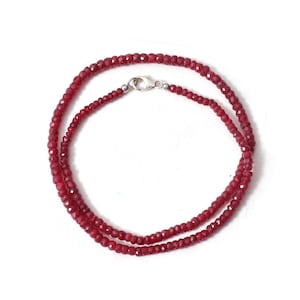 Può includere: Una collana composta da piccole perline sfaccettate di colore rosso intenso. Le perline sono infilate insieme, formando un doppio filo collegato da una chiusura color argento. La collana è esposta su sfondo bianco.