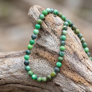 May include: A beaded necklace featuring round, green beads with varying shades of green and gray. The necklace is draped over a piece of weathered wood, showcasing the natural tones of the beads.