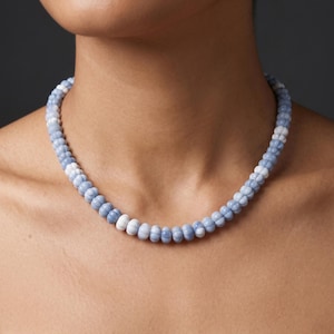 May include: A necklace made of light blue and white beads. The beads are small and round, and the necklace is worn around the neck. The background is a dark grey colour.