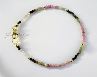 May include: A delicate bracelet made with small, colorful gemstones. The bracelet features a gradient of pink, green, and black gemstones, with a gold clasp. The bracelet is approximately 7 inches in length.
