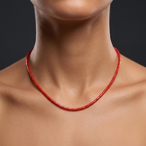 Peut inclure: Un collier composé de petites perles cylindriques d'un rouge éclatant enfilées sur un seul brin. Les perles sont uniformes en forme et en taille, créant une ligne circulaire lisse et continue autour du cou. La couleur est un rouge vif et saturé.