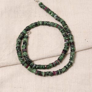 May include: A necklace made of small, round, green and maroon beads. The beads are strung together to form a necklace with a silver clasp. The necklace is displayed on a beige fabric surface.