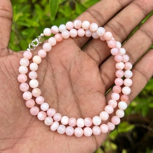 Puede incluir: Un delicado collar compuesto por pequeñas cuentas redondas de color rosa. Las cuentas están ensartadas juntas, formando una doble hebra. El collar se asegura con un cierre plateado. El fondo es una vista borrosa de follaje verde.