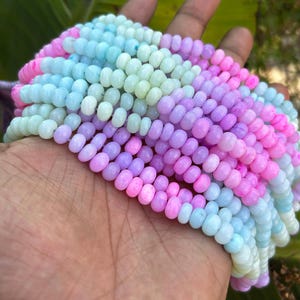 Puede incluir: Una hebra de cuentas de piedras preciosas de color pastel. Las cuentas son una mezcla de azul claro, verde, morado y rosa. Las cuentas son redondas y tienen un acabado liso y pulido.