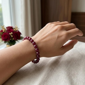 Puede incluir: Una pulsera elástica compuesta por cuentas redondas de piedra preciosa de color rojo oscuro con facetas. Las cuentas tienen una superficie pulida y reflectante, ensartadas en un bucle continuo que crea un diseño circular uniforme.