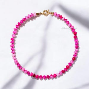 Puede incluir: Un collar de cuentas rosas vibrantes con un cierre dorado. El collar presenta una variedad de tonos rosas, desde claros hasta profundos, creando un efecto degradado. Las cuentas son pequeñas y redondas, ensartadas para formar un collar tipo gargantilla.