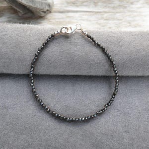 May include: A delicate bracelet featuring small, dark gray beads strung together. The bracelet is secured with a silver-colored clasp. The beads have a faceted appearance, reflecting light. The jewelry is displayed on a gray surface.