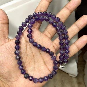 May include: A necklace made of round, polished amethyst beads. The beads are a deep purple color and strung together. The necklace is held in a hand, and the clasp is silver. The necklace is a jewelry item.