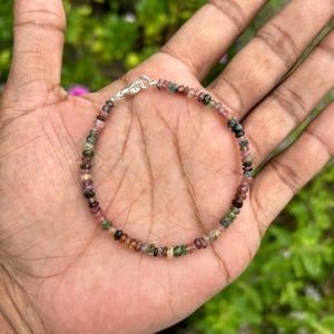 May include: A bracelet made of small, multi-colored gemstones. The bracelet is made of pink, green, and black gemstones. The bracelet is on a person's hand.