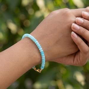 Op de afbeelding: Een lichtblauwe kralenarmband met een gouden sluiting. De armband is gemaakt van kleine, ronde kralen in verschillende tinten lichtblauw. De sluiting is goudkleurig. De armband wordt om een pols gedragen.