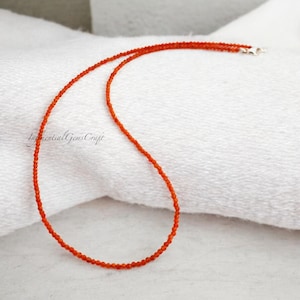 Peut inclure: Un collier délicat de perles orange avec un fermoir argenté. Le collier est présenté sur une surface blanche. Les perles sont petites et espacées uniformément, créant un design simple mais élégant. C'est un bijou.