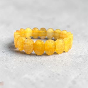 Könnte beinhalten: Ein Armband mit ovalen, gelben Perlen. Die Perlen sind von einem durchscheinenden, goldgelben Farbton und sind zu einem vollständigen Kreis aufgereiht. Das Armband ist vor einem neutralen grauen Hintergrund platziert.