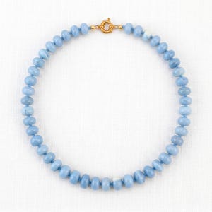 May include: A light blue beaded necklace with a gold-coloured clasp. The necklace features round, light blue beads strung together. The clasp has a small, ornate design. The necklace is displayed on a white surface.