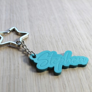 May include: A teal-coloured keychain with the name "Stephen" in a cursive font. It features a star-shaped silver metal ring and chain. The background is a light wood grain.