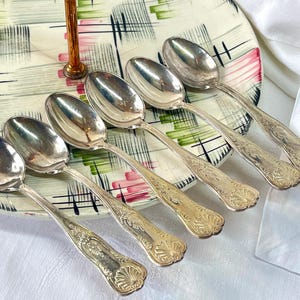 May include: A set of six silver-toned metal spoons with ornate embossed shell patterns on the handles. The spoons are arranged in a diagonal row across a round serving tray featuring a white surface with green and pink abstract brushstroke designs.