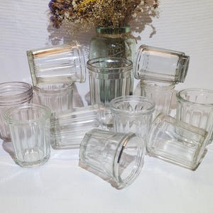 Puede incluir: Una colección de vasos de vidrio transparente y acanalado de varios tamaños, algunos tumbados de lado. Los vasos están dispuestos sobre una superficie blanca, con un jarrón de flores secas en el fondo.