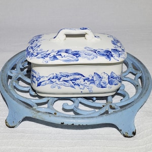 May include: A white ceramic butter dish with a blue floral design and a handle, resting on a light blue cast iron trivet. The trivet has a decorative, openwork design and three feet. The butter dish is rectangular with a matching lid.