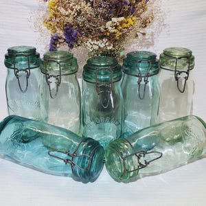 Op de afbeelding: Een verzameling vintage blauwgroene glazen potten met scharnierende deksels en metalen sluitingen. De potten zijn in verschillende maten en vormen, sommige rechtop en sommige liggend. Een boeket droogbloemen staat op de achtergrond.