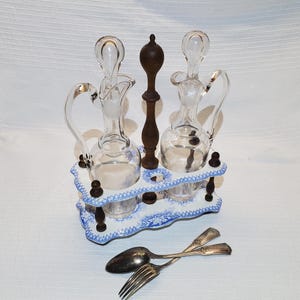 May include: A vintage cruet set featuring two clear glass bottles, a dark wooden central support, and a blue and white ceramic base. Two silver spoons and a fork are in the foreground. The set is designed for oil and vinegar.