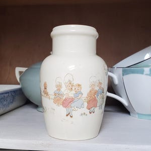 May include: A cream-colored ceramic vase featuring a decorative illustration of several children wearing white bonnets and colorful clothing sitting on a fence, with a small dog nearby. The vase has a smooth, rounded body and a flared rim at the top.