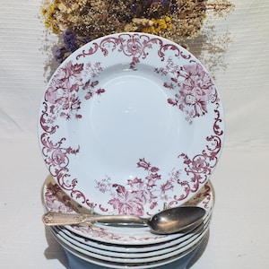 May include: A stack of white ceramic soup bowls featuring a deep red floral transferware pattern along the wide rims. A tarnished silver-plated spoon with an ornate handle rests horizontally across the top bowl of the stack.