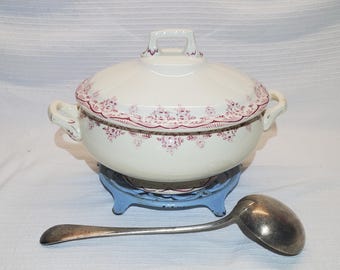 May include: A white ceramic soup tureen with a lid, adorned with a burgundy floral pattern. It has two side handles and a handle on the lid. A silver ladle rests beside the tureen, which sits on a light blue metal stand.