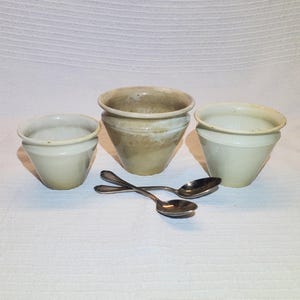 Peut inclure: Trois petits pots en céramique blanc cassé de forme légèrement évasée. Deux cuillères en argent sont croisées devant les pots. Les pots ont un aspect rustique et fait à la main, adaptés aux petites plantes ou à un usage décoratif.