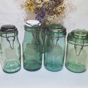 Peut inclure: Quatre bocaux en verre vert vintage avec couvercles à charnière et fermoirs métalliques. Les bocaux varient en forme et en taille, avec le texte "L'IDEALE" visible sur deux d'entre eux. Un bouquet de fleurs séchées est en arrière-plan.
