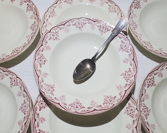 May include: A collection of white soup bowls with a burgundy floral pattern around the rim. A silver spoon rests in one of the bowls. The bowls are arranged on a white surface, creating a classic, vintage aesthetic.