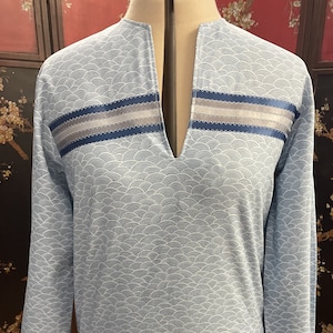 May include: A light blue long-sleeved shirt with a white wave pattern. The shirt has a V-neck and a blue and gray ribbon trim around the neckline.
