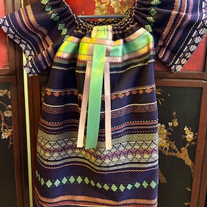 Puede incluir: Un vestido de rayas azul oscuro y marrón con una cinta verde y rosa atada en el escote. El vestido tiene un patrón geométrico y está hecho de una tela tejida.