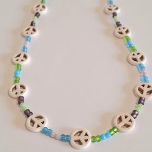 Può includere: Una collana di perline con ciondoli circolari bianchi a forma di simbolo della pace, intervallati da piccole perline di vetro traslucido nei toni del verde lime, azzurro cielo e viola tenue, disposti a forma di U.