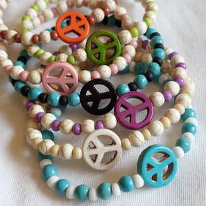 Könnte beinhalten: Ein Set aus sieben bunten Armbändern mit Peace-Zeichen-Anhängern. Die Armbänder bestehen aus weißen Perlen, die mit verschiedenen farbigen Perlen durchsetzt sind. Die Peace-Zeichen-Anhänger sind aus Holz und in verschiedenen Farben bemalt.