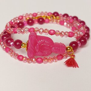 Può includere: Un braccialetto di perline rosa con un ciondolo a forma di Buddha rosa e un fiocco rosso.