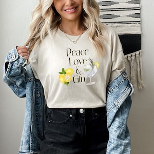 Op de afbeelding: Een beige T-shirt met de tekst "Peace Love & Gin" en een afbeelding van een gin tonic met citroenen.