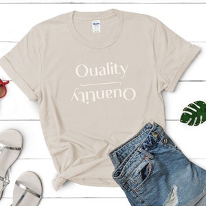 May include: A beige t-shirt with the text "Quality over Quantity" printed in white on the front. The shirt is folded and displayed on a white wooden surface with a pair of silver sandals, a pair of red sunglasses, and a green leaf keychain.