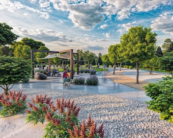 May include: A park setting with a wooden pergola, a gravel path, and lush green trees. People are walking through the park, and there are benches and a small dog.