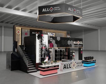 May include: A two-story trade show booth for ALL Armor Life Lab featuring black and white branding, a hexagonal hanging sign, a staircase, product display counters with illuminated bases, and large vertical banners with graphic imagery and text.