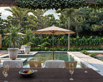 May include: Outdoor dining scene with a dark wood table set with champagne flutes and a bowl of fruit. A beige umbrella shades a pool surrounded by lush greenery and palm trees. The scene evokes a sense of relaxation and luxury.