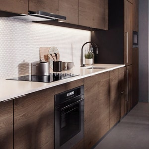 Puede incluir: Una cocina moderna con gabinetes de madera oscura y una encimera blanca. La cocina cuenta con un horno negro, una placa de cocina y una campana extractora. Una olla de acero inoxidable y utensilios están en la placa de cocina. El salpicadero es blanco con azulejos pequeños y redondos.