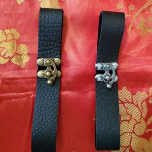 May include: Two black leather straps with metal clasps. One clasp is brass-colored, the other is silver-colored. The straps are rectangular and have a textured surface. The background is red with a gold floral pattern.