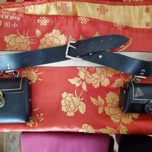 Puede incluir: Un cinturón de cuero negro con hebilla plateada y dos bolsos de cuero negro a juego con cierres ornamentados. Los artículos se exhiben sobre una tela estampada floral roja y dorada.